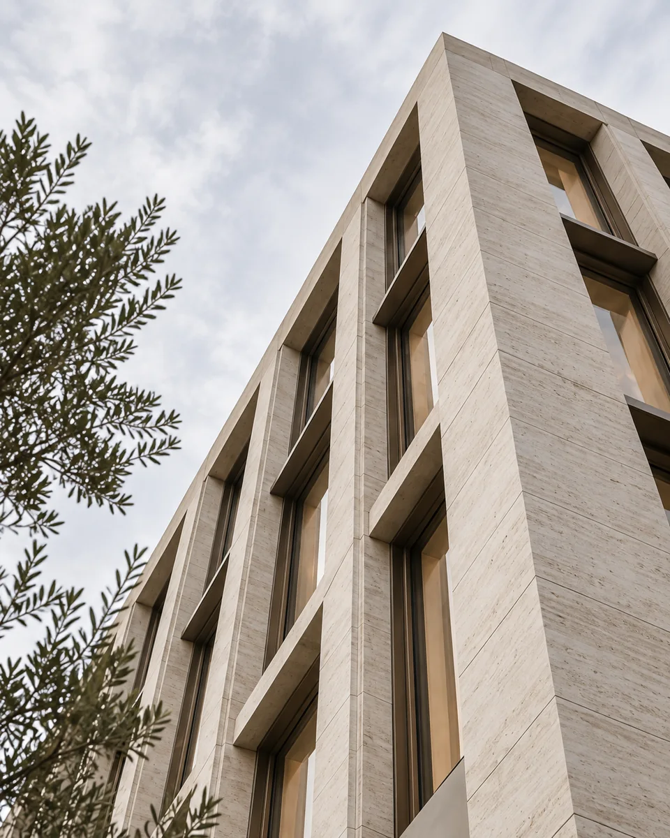 A pale limestone facade with recessed bronze-framed windows and olive foliage.